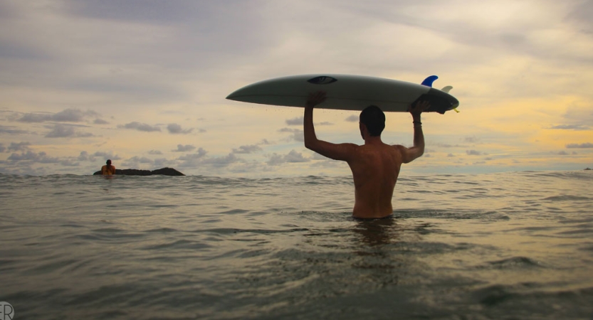 a person standing in waist-deep water balances a surfboard on their head and looks out at the water